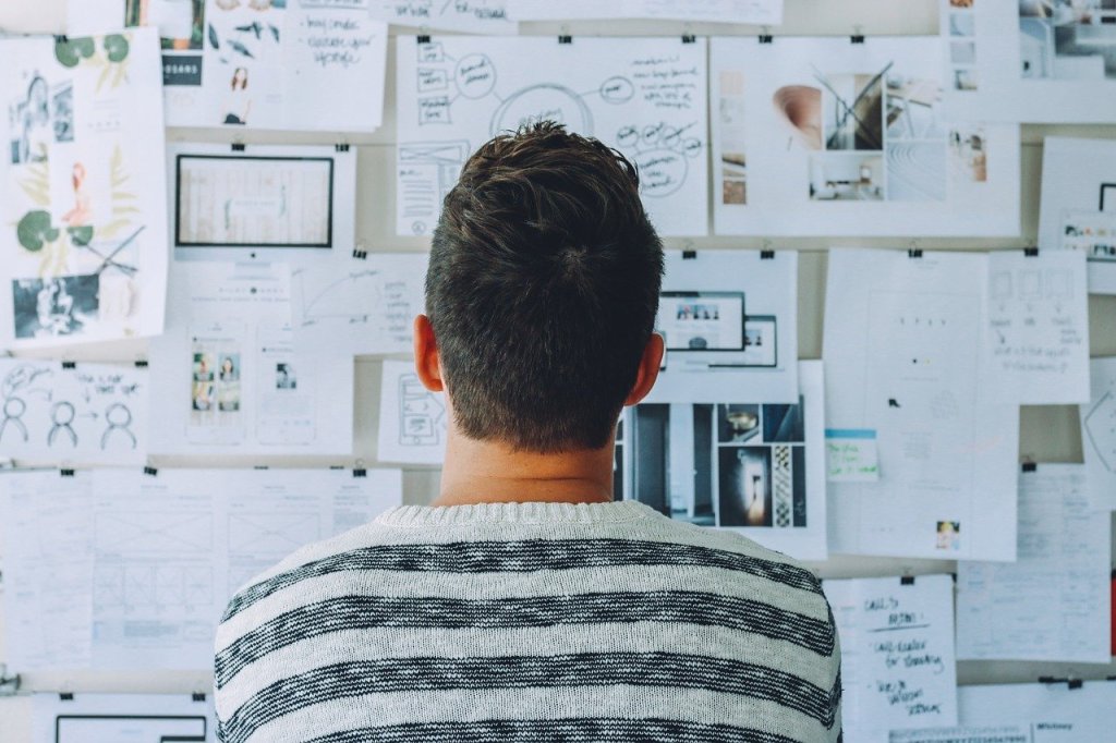 A man in a stripped shirt looking at a full wall of pinned papers with various graphs and notes on them. The man is in focus while the wall is slightly blurred.