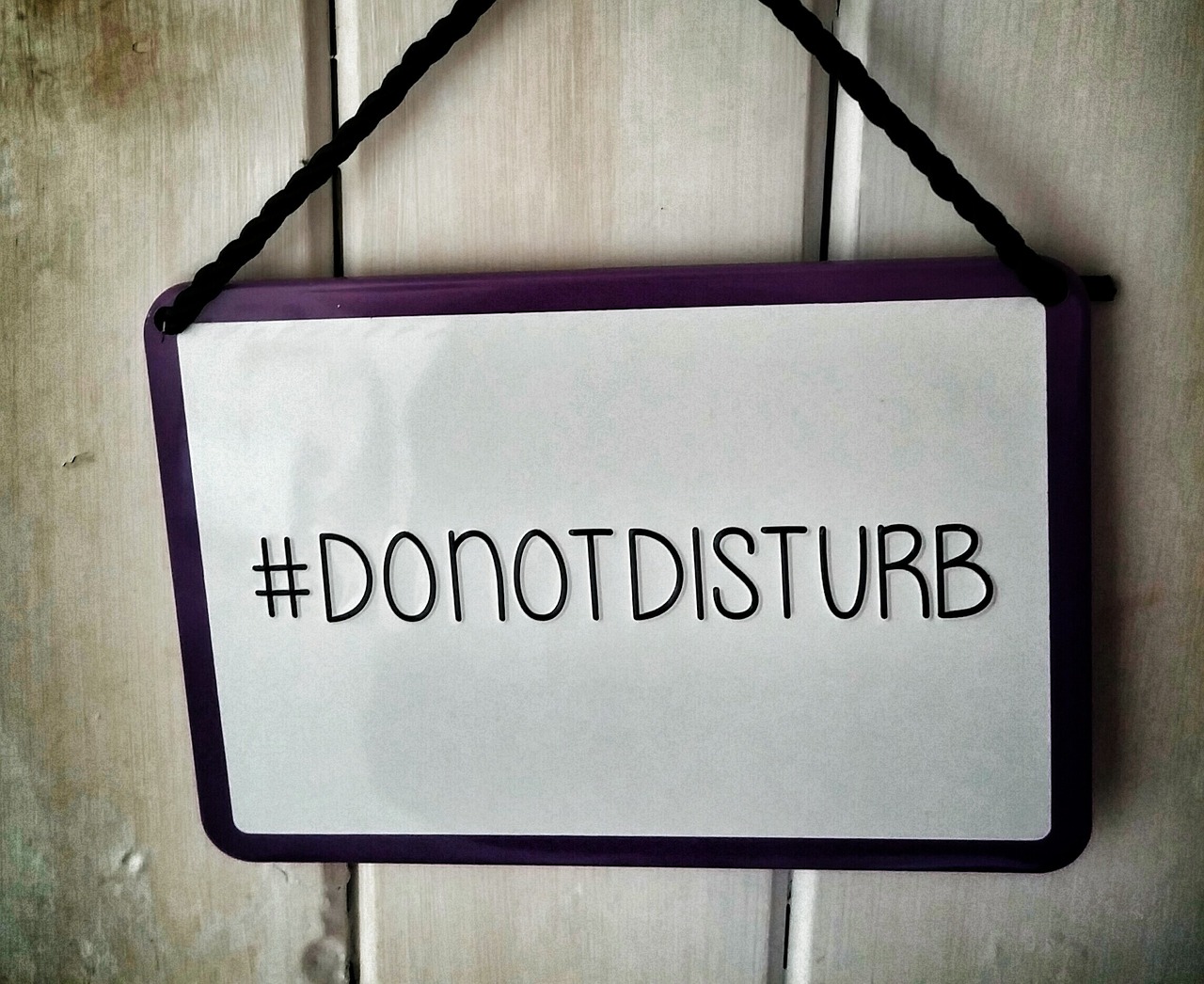 A white sign with a black border against a wooden wall. The sign reads in black letters #DONOTDISTURB